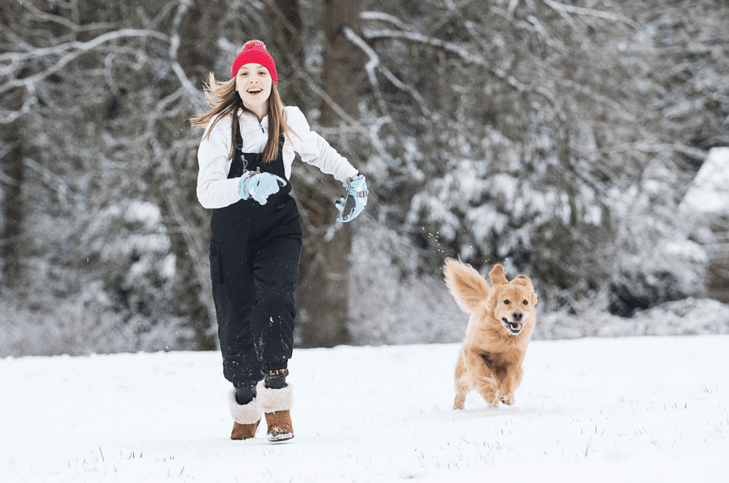 Smiling girl in winter clothes running through fresh snow beside a happy golden dog.