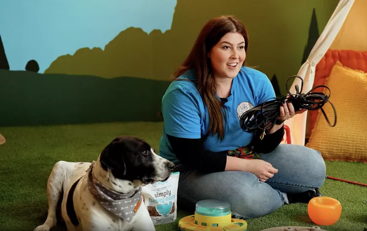 Dog trainer sitting on the floor beside a spotted dog, holding a neatly coiled long line leash with treats and toys nearby.